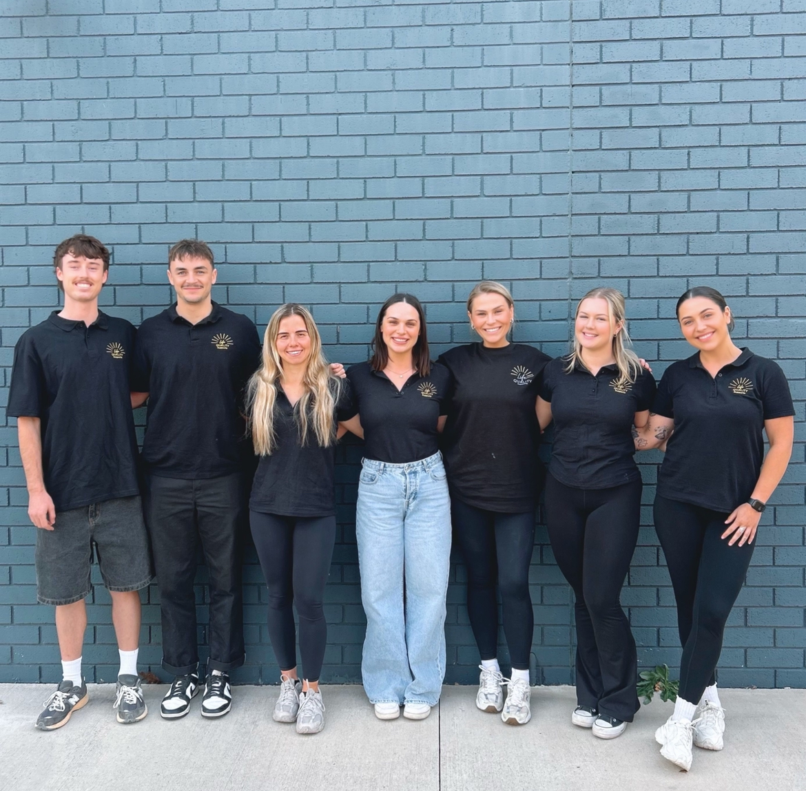 A diverse team of support workers posing together outside against a blue brick wall, all wearing black polo shirts with the company logo.